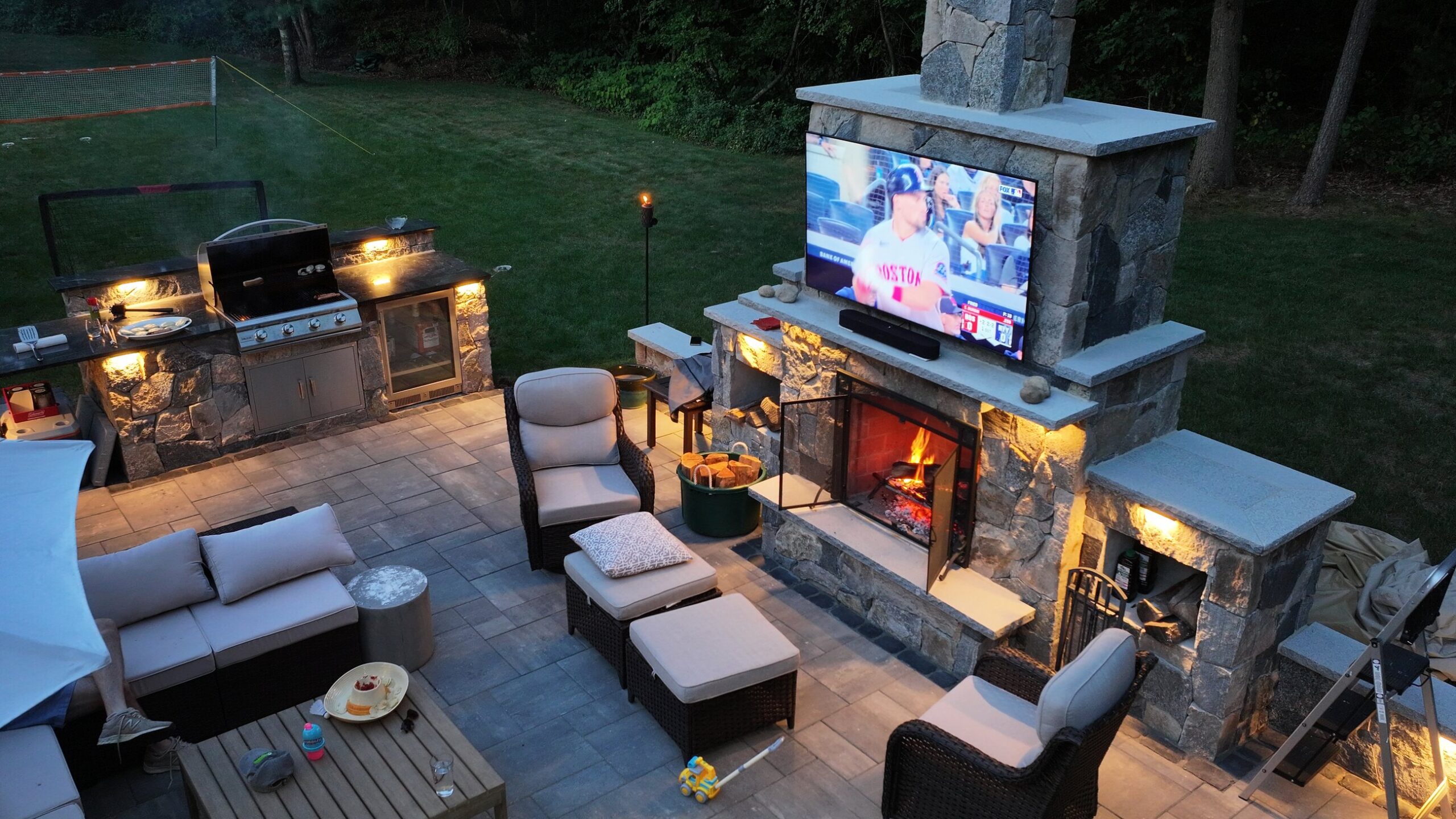 Duxbury outdoor living space with masonry fireplace and outdoor kitchen illuminated at dusk